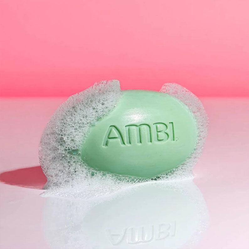 AMBI green oval complexion cleansing bar with embossed logo and foamy lather on glossy surface against pink background