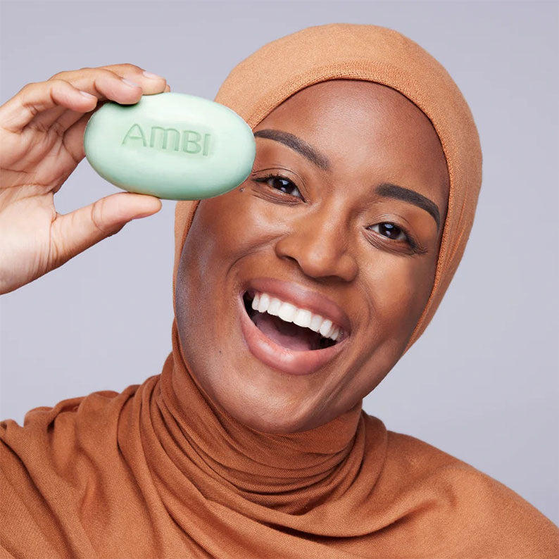 Smiling woman in brown hijab holding AMBI green complexion cleansing bar near her face for gentle skincare