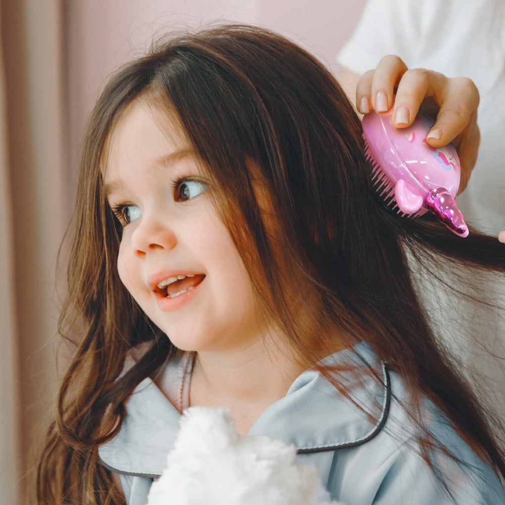 Child smiling while adult uses pink unicorn detangling brush on long brown hair for gentle grooming